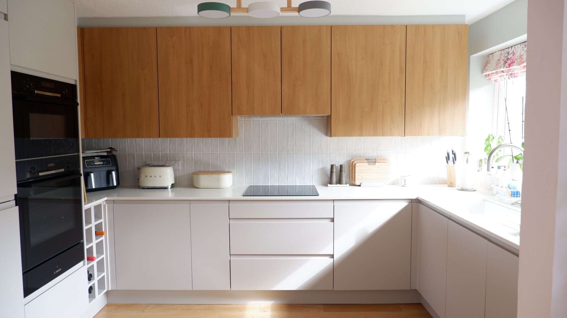 A modern, handleless kitchen with light grey and warm wood cupboards. Natural light beams in through the window.