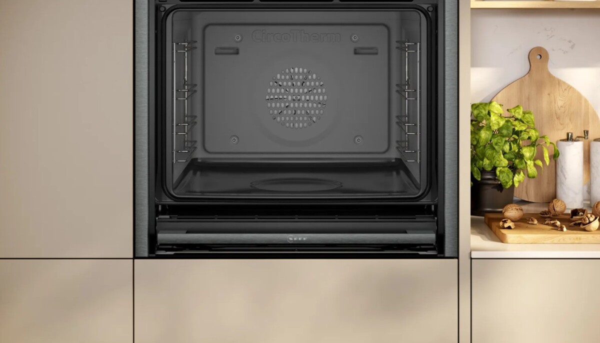 An integrated Neff slide and hide oven in a neutral kitchen with a plant and chopping boards on the counter
